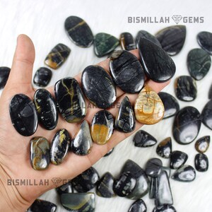 May include: A collection of black and gold gemstone cabochons, some with a teardrop shape, others oval, and some with a more irregular shape. The stones are arranged on a hand and a white surface. Bismillah Gems is printed on the image.