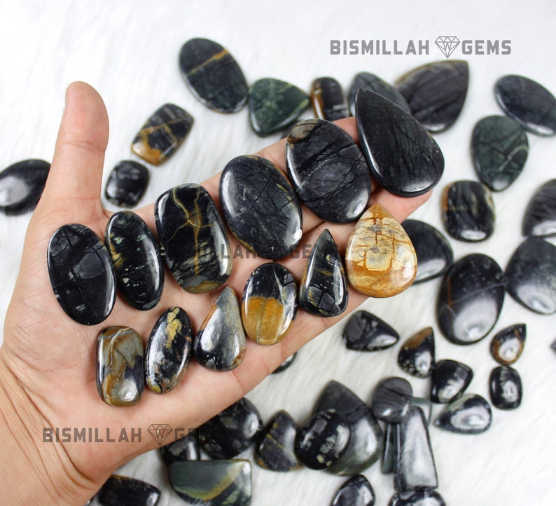 May include: A collection of black and gold gemstone cabochons, some with a teardrop shape, others oval, and some with a more irregular shape. The stones are arranged on a hand and a white surface. Bismillah Gems is printed on the image.