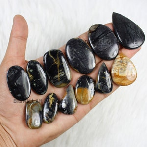 May include: A collection of 11 polished black and brown gemstone cabochons in various shapes, including oval, teardrop, and pear. The stones have a unique marbled pattern with black, brown, and white colours.