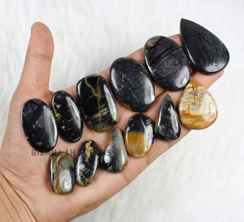May include: A collection of 11 polished black and brown gemstone cabochons in various shapes, including oval, teardrop, and pear. The stones have a unique marbled pattern with black, brown, and white colours.