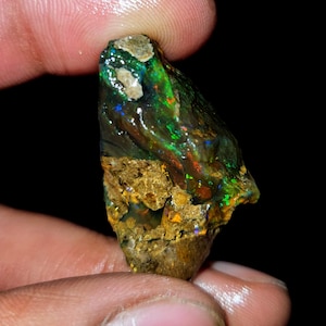 Può includere: Una pietra preziosa di opale grezza, con una base scura e terrosa. L'opale mostra vibranti lampi di verde, blu e rosso, creando un gioco di colori. La pietra è tenuta tra le dita su uno sfondo nero, evidenziando la sua bellezza naturale.