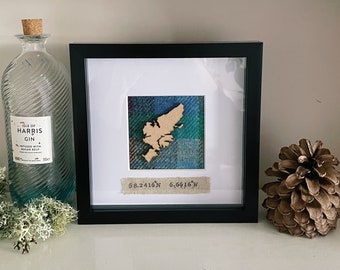 May include: A square black frame containing a wooden cutout of a landmass mounted on blue and green plaid fabric. Below the cutout, a beige fabric strip displays the printed coordinates 58.2416 N 6.6616 W. The frame is set against a white background.