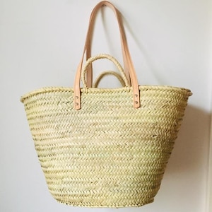 Könnte beinhalten: Eine große, naturgeflochtene Strohtasche mit zwei langen Ledergriffen. Die Griffe haben Metallnieten und sind oben an der Tasche befestigt.