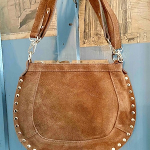 Può includere: Borsa a tracolla in pelle scamosciata marrone con base curva e tracolla lunga. La borsa è decorata con piccole borchie argentate lungo i bordi. La borsa ha un aspetto morbido e strutturato e uno stile vintage.