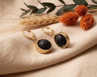 May include: A pair of gold-toned leverback earrings featuring a circular frame that holds a porous, spherical black lava stone. The metal components have a smooth, polished finish, while the dark stones provide a textured contrast.