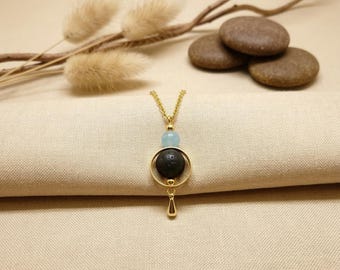 May include: A gold-tone chain necklace featuring a pendant with a small light blue bead, a circular gold frame containing a textured black porous stone, and a small gold teardrop-shaped charm hanging from the bottom of the circular frame.