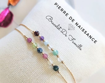 May include: Three delicate gold chain bracelets with colourful gemstone beads. The bracelets are displayed on a white fabric surface. The text "Pierre de Naissance Bracelet De Famille" is visible above the bracelets.
