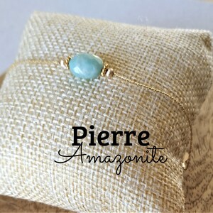 May include: A delicate gold chain bracelet with a single light blue amazonite gemstone bead. The bracelet is displayed on a beige fabric surface with the text "Pierre Amazonite".