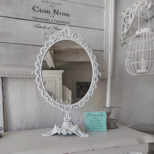 Peut inclure: Un miroir ovale blanc orné avec un support décoratif. Le miroir est entouré d'un cadre détaillé en forme de fleur. Un petit panneau turquoise avec du texte est visible. Une figurine de cerf blanche et un panier tressé sont également sur la table.