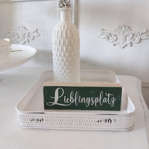 Può includere: Un vassoio decorativo bianco con bordo traforato contiene un vaso bianco strutturato con fiori secchi e un cartello di legno verde con la scritta "Lieblingsplatz" in corsivo. La scena è ambientata su uno sfondo bianco.