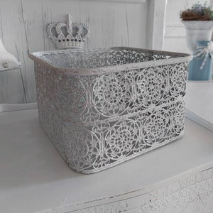 Könnte beinhalten: Ein quadratischer, durchbrochener Metallkorb in einem weißen Used-Look. Der Korb weist ein sich wiederholendes kreisförmiges Blumenmuster auf und steht auf einer weißen Oberfläche. Im Hintergrund ist eine dekorative Krone zu sehen, die auf eine Vintage- oder Shabby-Chic-Ästhetik hindeutet.