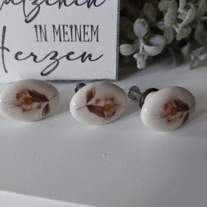 Peut inclure: Trois boutons de tiroir en céramique blanche avec des motifs floraux marrons. Les boutons ont une fixation à vis en métal.