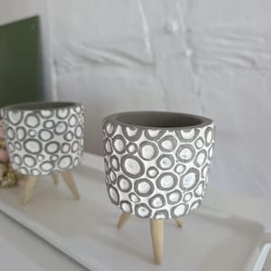 Puede incluir: Dos maceteros cilíndricos con un patrón texturizado de círculos superpuestos en gris y blanco. Cada macetero se apoya sobre tres patas de madera de color claro, proporcionando una base estable. El diseño es consistente en ambos objetos.
