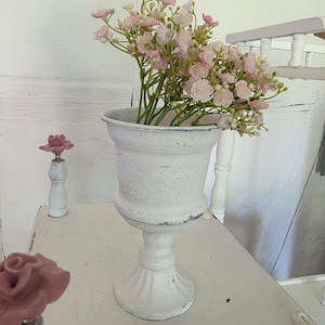 Puede incluir: Un florero de pedestal blanco con un acabado texturizado y desgastado que contiene un ramo de flores artificiales de color rosa claro. El florero presenta un borde ancho y acampanado y una base circular resistente con marcas de desgaste.