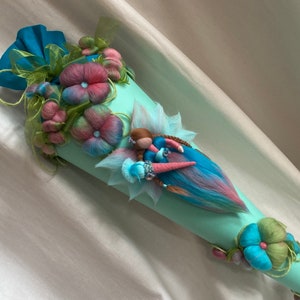 Könnte beinhalten: Ein hellblauer Kegel, der mit Filzblumen und einer Feenpuppe verziert ist. Die Fee hat rosa und blaue Haare, ein blaues Kleid und einen rosa und blauen Schwanz.