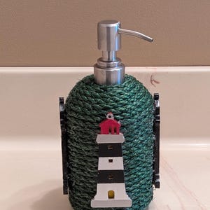 May include: A decorative soap dispenser with a green rope-wrapped body and a silver pump. The dispenser is adorned with a lighthouse design in red, white, and black, adding a nautical touch to bathroom decor.