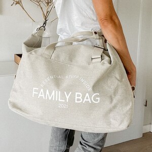May include: A beige canvas tote bag with the words "FAMILY BAG" printed in white. The text "ESSENTIAL STUFF INSIDE" is arched above the main text, and "2021" is printed below. The bag has a shoulder strap and handles.