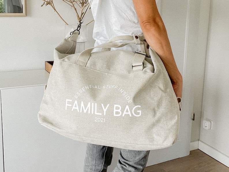 May include: A beige canvas tote bag with the words "FAMILY BAG" printed in white. The text "ESSENTIAL STUFF INSIDE" is arched above the main text, and "2021" is printed below. The bag has a shoulder strap and handles.