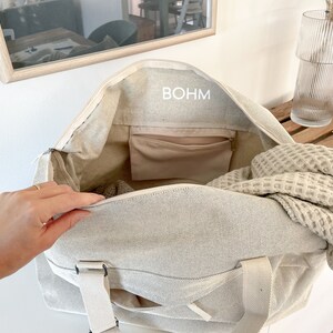 May include: Beige canvas tote bag with the word "BOHM" printed on the inside. The bag has a zippered closure, a small interior pocket, and cream-colored straps. A waffle-weave blanket is partially visible inside the bag.