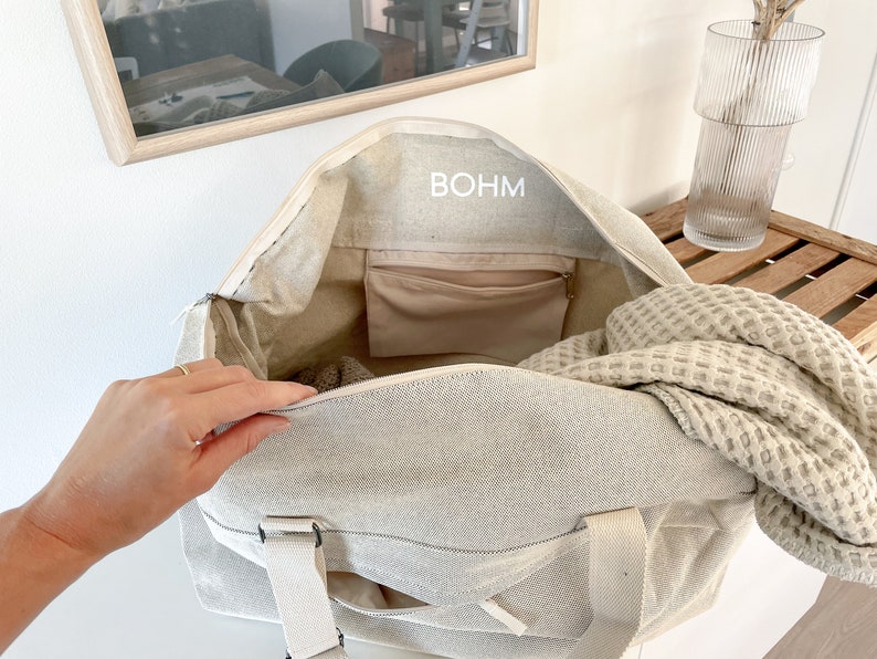 May include: Beige canvas tote bag with the word "BOHM" printed on the inside. The bag has a zippered closure, a small interior pocket, and cream-colored straps. A waffle-weave blanket is partially visible inside the bag.