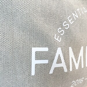 May include: Close-up of a beige tote bag with a textured, dotted pattern. The words "ESSENTIAL STYLE" and "FAMILY" are printed in white. The year range "2016-2018-2023" is also visible. The bag appears to be made of a durable fabric.