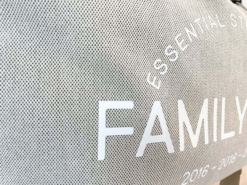 May include: Close-up of a beige tote bag with a textured, dotted pattern. The words "ESSENTIAL STYLE" and "FAMILY" are printed in white. The year range "2016-2018-2023" is also visible. The bag appears to be made of a durable fabric.