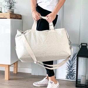 May include: A large, beige canvas tote bag with a shoulder strap and top handles. The bag is rectangular and appears to be made of a textured fabric. The bag is being held by a person wearing black pants and white sneakers.