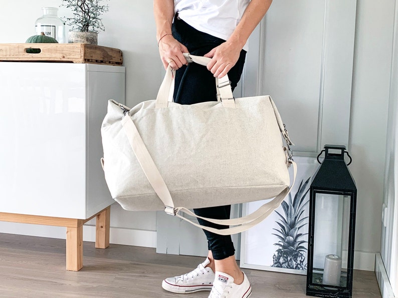 May include: A large, beige canvas tote bag with a shoulder strap and top handles. The bag is rectangular and appears to be made of a textured fabric. The bag is being held by a person wearing black pants and white sneakers.