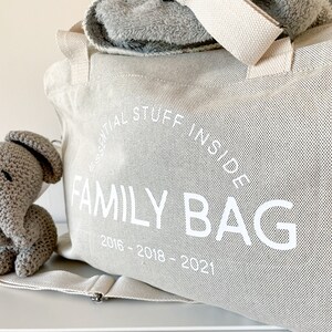 May include: A light beige canvas family bag with the text "ESSENTIAL STUFF INSIDE FAMILY BAG 2016-2018-2021" in white. A gray crocheted elephant toy sits next to the bag. A gray blanket is visible inside the bag.