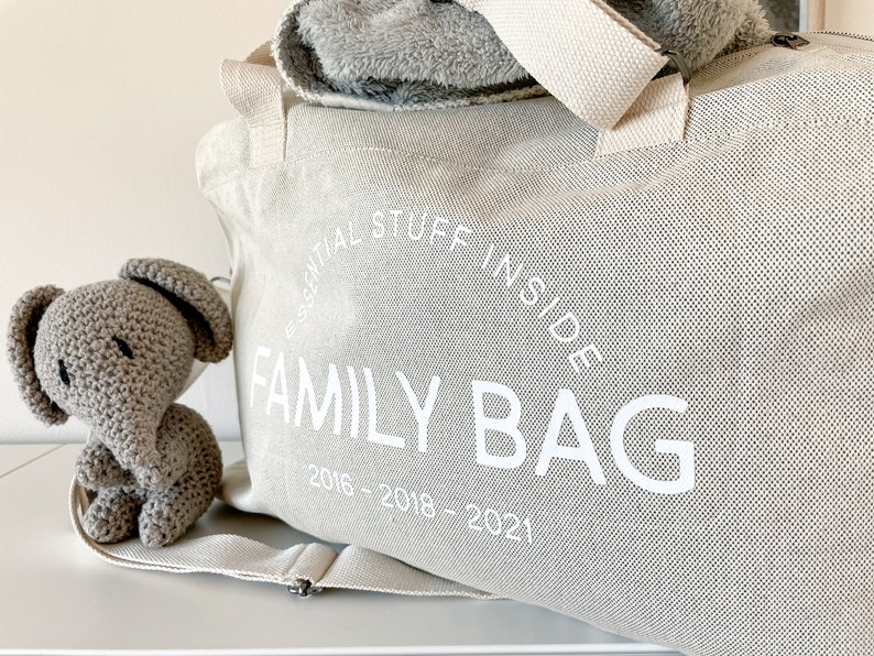 May include: A light beige canvas family bag with the text "ESSENTIAL STUFF INSIDE FAMILY BAG 2016-2018-2021" in white. A gray crocheted elephant toy sits next to the bag. A gray blanket is visible inside the bag.