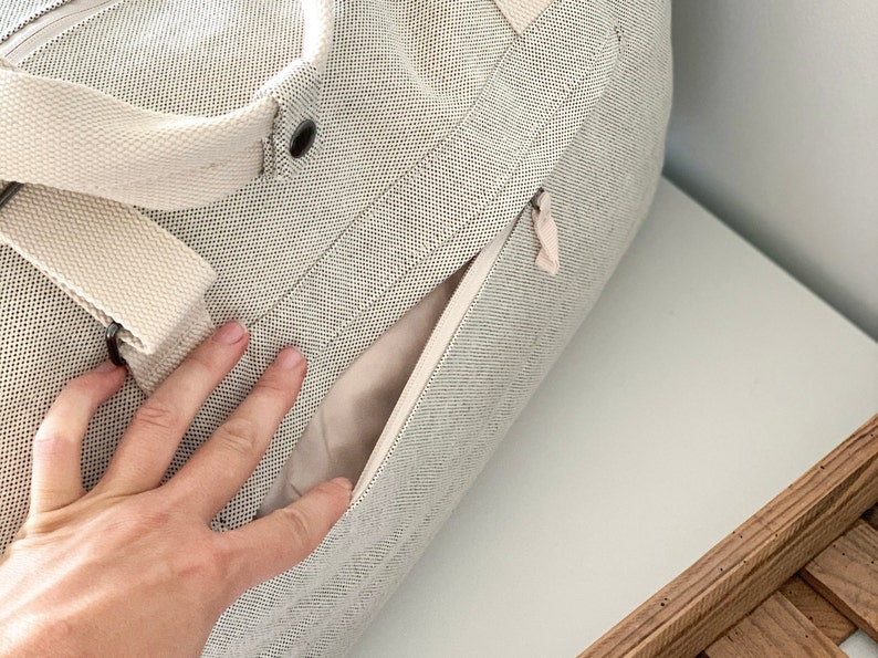 May include: A light gray canvas tote bag with a cream-colored strap and a side zipper pocket. The bag has a textured weave and a neutral color palette, suitable for various uses.
