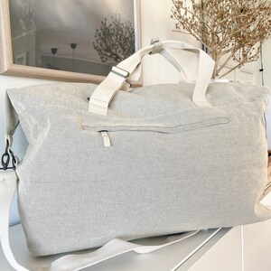May include: A large, light gray canvas tote bag with a zippered pocket and cream-colored straps. The bag has a shoulder strap and two top handles. The bag is sitting on a white surface.