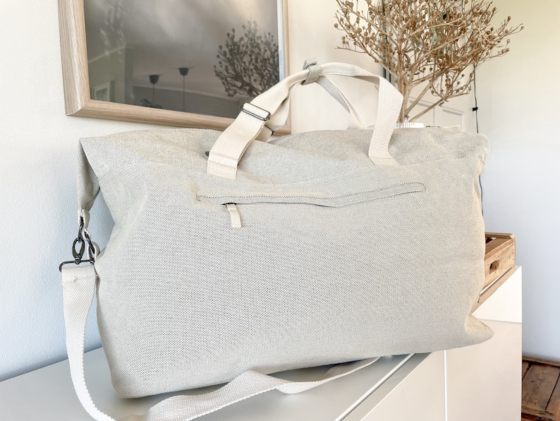 May include: A large, light gray canvas tote bag with a zippered pocket and cream-colored straps. The bag has a shoulder strap and two top handles. The bag is sitting on a white surface.