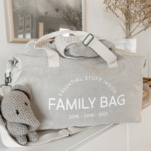May include: A light gray canvas family bag with the text "ESSENTIAL STUFF INSIDE FAMILY BAG 2016-2018-2021" in white. A small gray crocheted elephant is attached to the bag. A white strap and a rolled-up towel are on top.