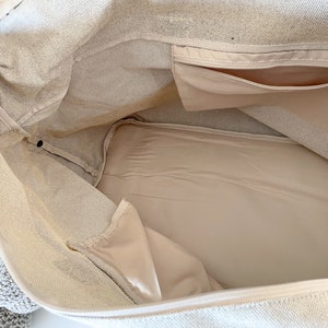 May include: Beige canvas tote bag interior with a zippered pocket and a smaller pocket. The inside lining is a lighter beige color. The bag is open, showing the inside structure and design.