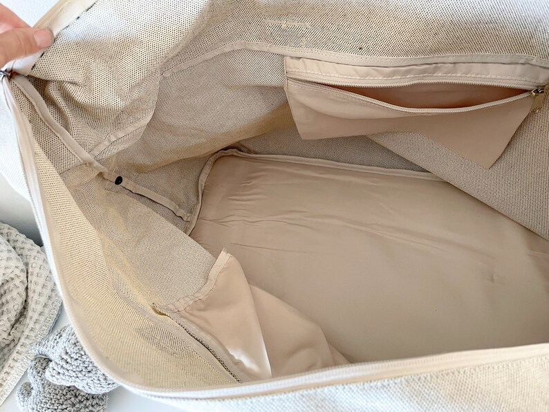 May include: Beige canvas tote bag interior with a zippered pocket and a smaller pocket. The inside lining is a lighter beige color. The bag is open, showing the inside structure and design.