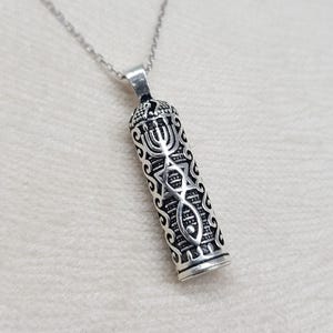 Peut inclure: Collier pendentif argenté avec un pendentif cylindrique orné de symboles religieux. Le pendentif présente une étoile de David, un menorah et d'autres motifs détaillés. La chaîne du collier est également argentée.