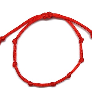 Pode incluir: Uma pulseira vermelha brilhante ajustável. A pulseira é feita de um cordão tecido e apresenta vários nós. Possui um fecho de nó deslizante para um ajuste personalizado. O design é simples e minimalista.