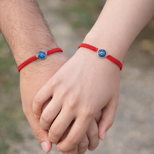 Puede incluir: Dos pulseras rojas tejidas, cada una con un dije circular plateado con un diseño de ojo azul y blanco. Las pulseras se usan en las muñecas, con las manos entrelazadas.