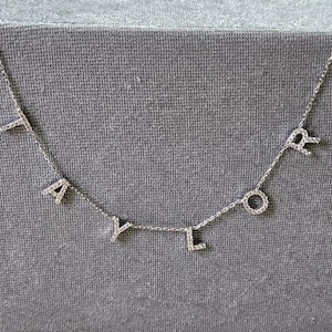 Puede incluir: Un collar de plata con la palabra "TAYLOR" escrita en pequeñas letras engastadas con diamantes.