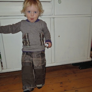 May include: A young child wearing a grey sweater with plaid patches and brown patchwork trousers stands in front of a white cabinet.