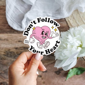 May include: A sticker with a pink heart character that has arms, legs, and a face. The sticker reads "Don't Follow Your Heart" in black text, with the Bible verse "Jer. 17:9" below the heart. The background includes a white flower and a white cloth.