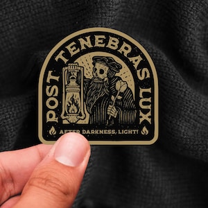 May include: A gold sticker with a black and white illustration of a skeleton holding a lantern and a flower. The text "POST TENEBRAS LUX" is written in a circle around the skeleton. The text "AFTER DARKNESS, LIGHT!" is written below the skeleton.