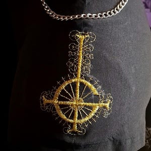 Könnte beinhalten: Ein schwarzer Rock mit einem goldenen, gestickten umgekehrten Kreuz, auch bekannt als umgekehrter Pentagramm, auf der Rückseite. Der Rock hat einen silbernen Kettengürtel.