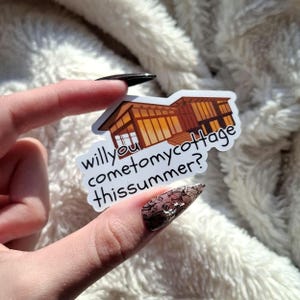Könnte beinhalten: Ein Aufkleber mit einem braunen Ferienhaus mit großen Fenstern und dem Text "will you come to my cottage this summer?" in einer handschriftlichen Schriftart. Der Aufkleber wird vor einem flauschigen weißen Hintergrund gehalten.
