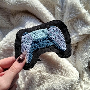 Könnte beinhalten: Gestickter Aufnäher mit einem Videospiel-Controller-Design. Der Controller ist in Blau- und Hellblautönen gehalten und auf einem schwarzen Stoffrücken aufgenäht. Der Aufnäher befindet sich vor einem weichen, weißen, strukturierten Hintergrund.
