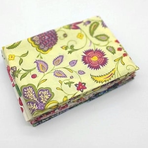 Può includere: Una pila di quadrati di tessuto floreale. Il tessuto presenta un motivo ripetuto di fiori e foglie viola, rossi e gialli su uno sfondo giallo pallido. Il tessuto è piegato ordinatamente, mostrando i bordi dei diversi strati.