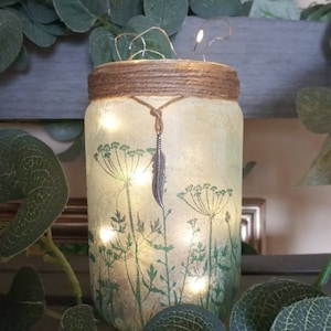 May include: A glass jar with rustic brown twine tied around the top. The jar is decorated with a green and white floral design and a silver feather charm. The jar is lit from within with small white lights.