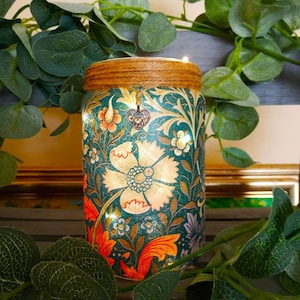 May include: A teal blue glass jar with a floral pattern and gold accents. The jar is decorated with a brown twine bow and a small silver heart charm. The jar is lit from within, creating a warm glow.