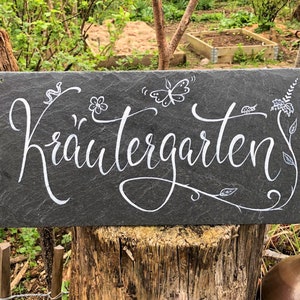 Op de afbeelding: Een zwart leisteenbord met witte letters die "Krautergarten" lezen. Het bord is versierd met kleine tekeningen van bloemen, vlinders en bladeren.